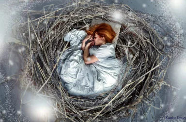 Digital artwork by Katelin Kinney showing a red-haired woman sleeping peacefully in a fetal position inside a large bird's nest made of dry branches and twigs, covered with a white blanket and resting her head on a small pillow, surrounded by soft glowing lights and a winter atmosphere.