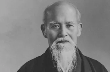 Historical portrait of Morihei Ueshiba, the founder whose teachings established the philosophy of aikido, showing his serene face with a long white beard and mustache against a neutral studio background.